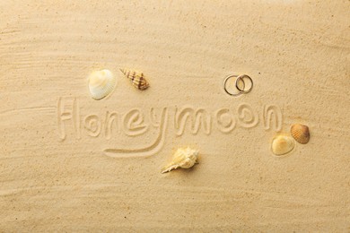 Photo of Word Honeymoon written on sand, two golden rings and seashells, flat lay