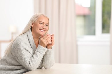 Portrait of beautiful older woman with cup of tea sitting at table indoors. Space for text