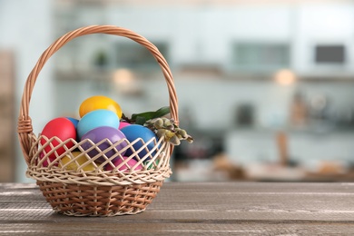 Image of Wicker basket with bright painted Easter eggs on wooden table indoors, space for text 