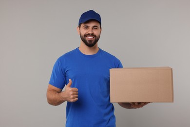 Photo of Happy young courier with parcel showing thumb up on grey background