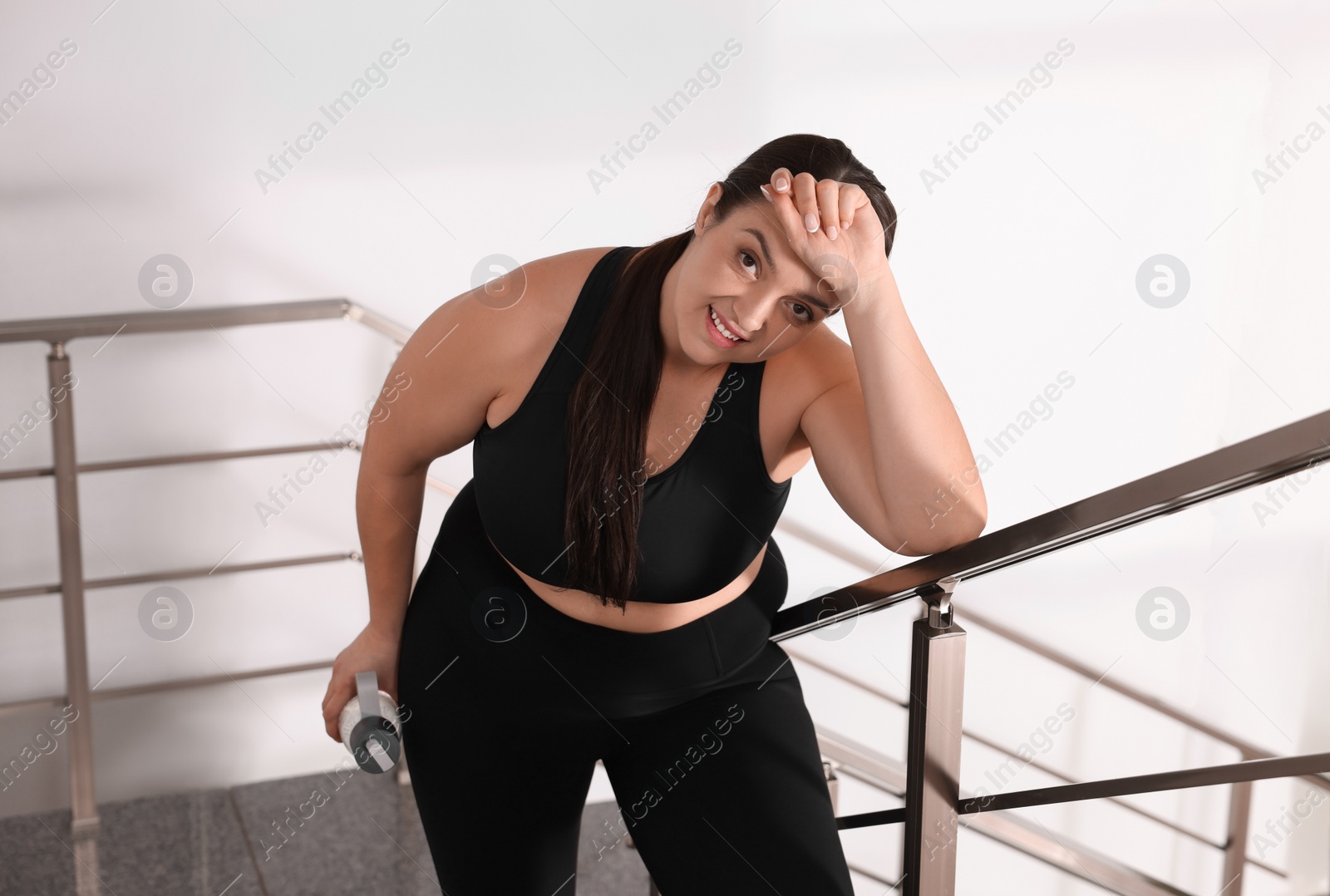 Photo of Tired overweight woman with bottle of water on stairs indoors