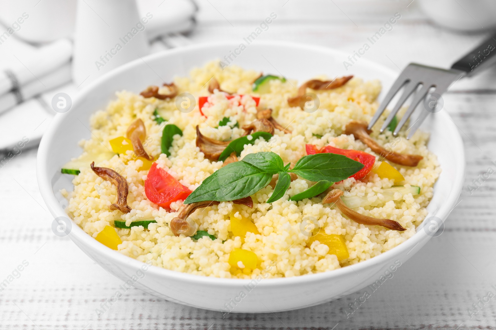 Photo of Bowl of delicious couscous with vegetables and basil served on white wooden table, closeup