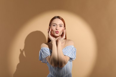 Photo of Beautiful young woman sending air kiss in sunlight on beige background