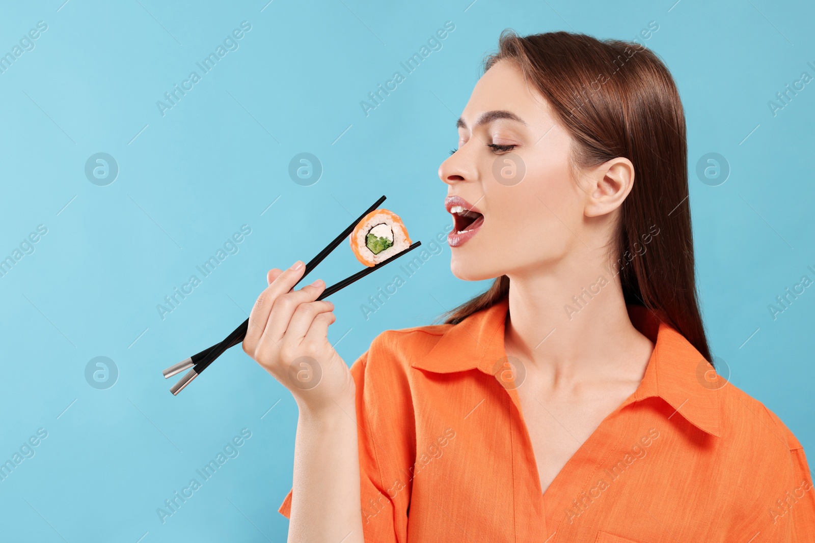 Photo of Beautiful young woman eating sushi roll with chopsticks on light blue background