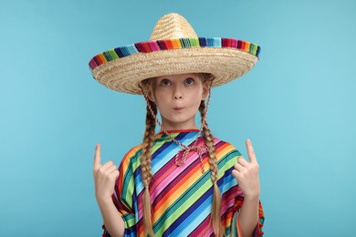 Cute girl in Mexican sombrero hat and poncho pointing at something on light blue background
