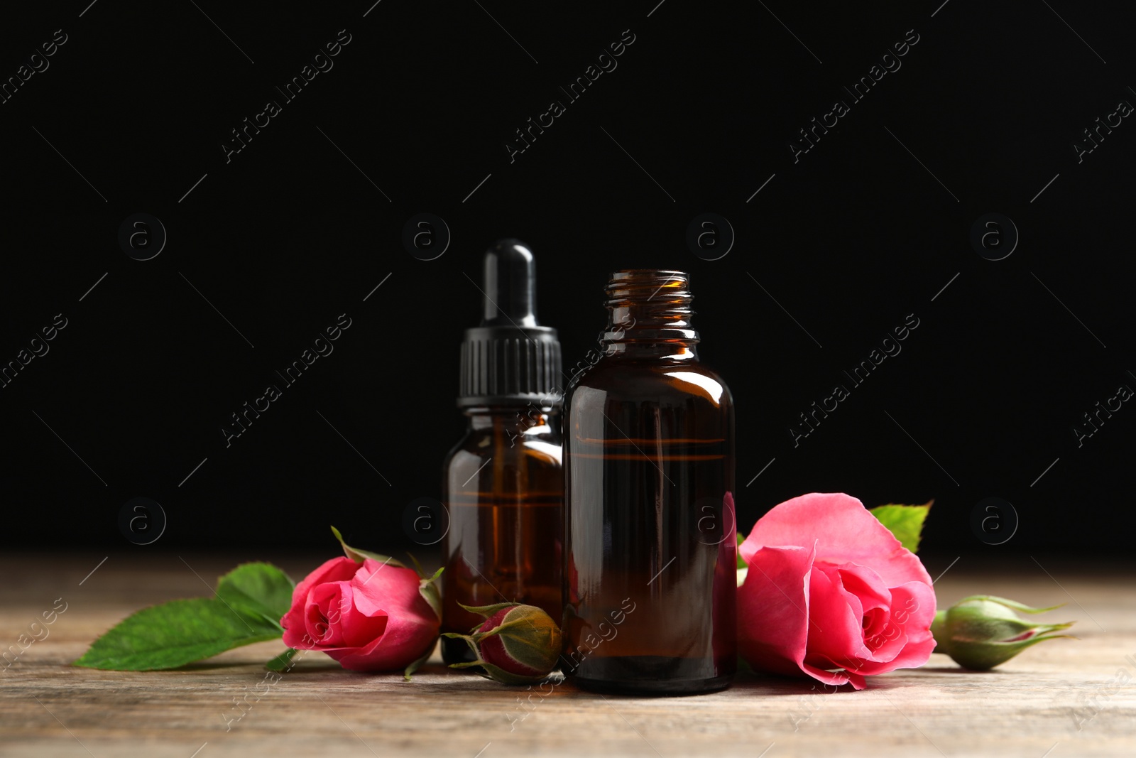 Photo of Bottles of rose essential oil and flowers on wooden table against black background