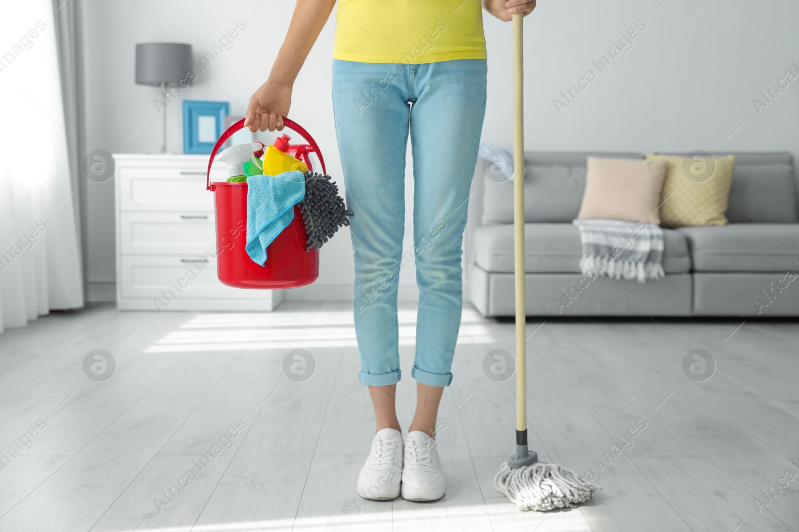 Photo of Woman holding bucket with different cleaning supplies and mop at home, closeup