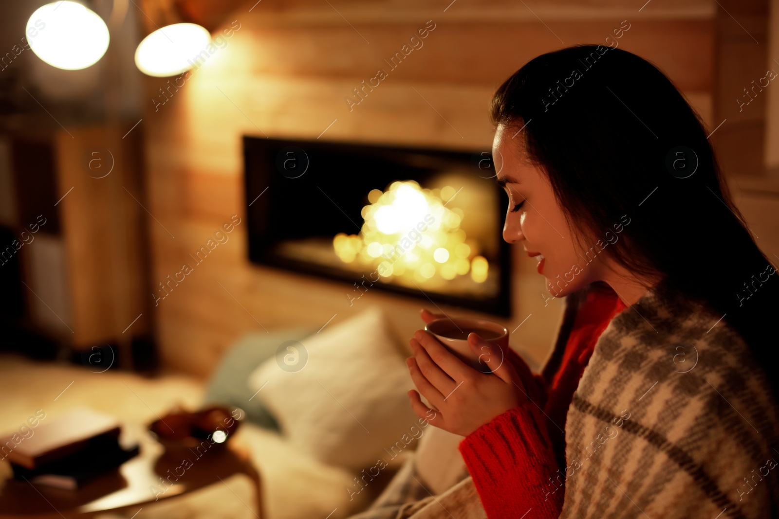 Photo of Young woman drinking coffee near decorative fireplace at home. Winter season