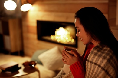 Photo of Young woman drinking coffee near decorative fireplace at home. Winter season