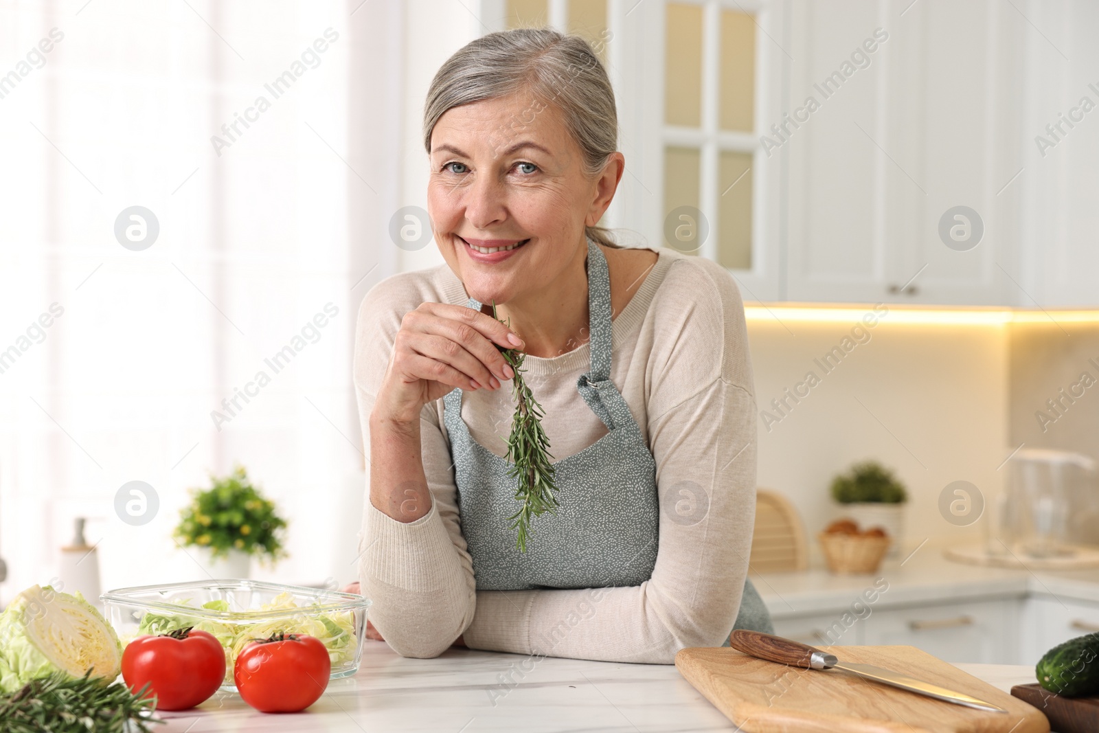Photo of Happy housewife with rosemary at white marble table in kitchen. Cooking process