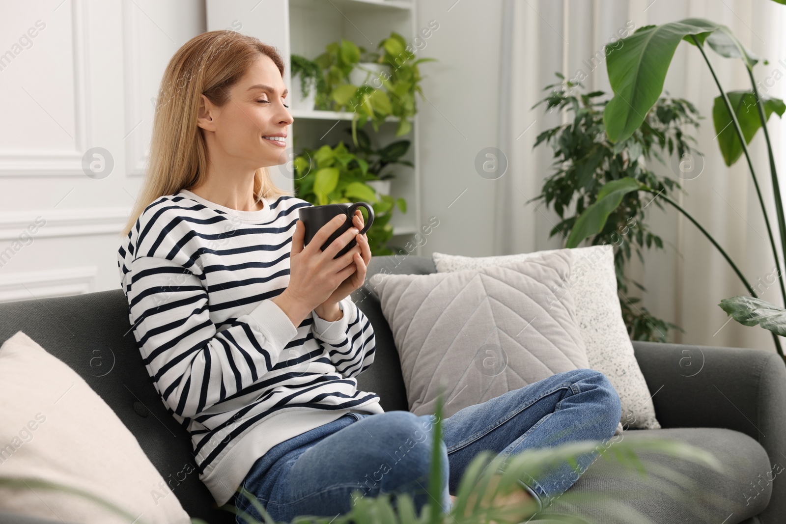 Photo of Woman with cup of drink sitting on sofa surrounded by beautiful potted houseplants at home