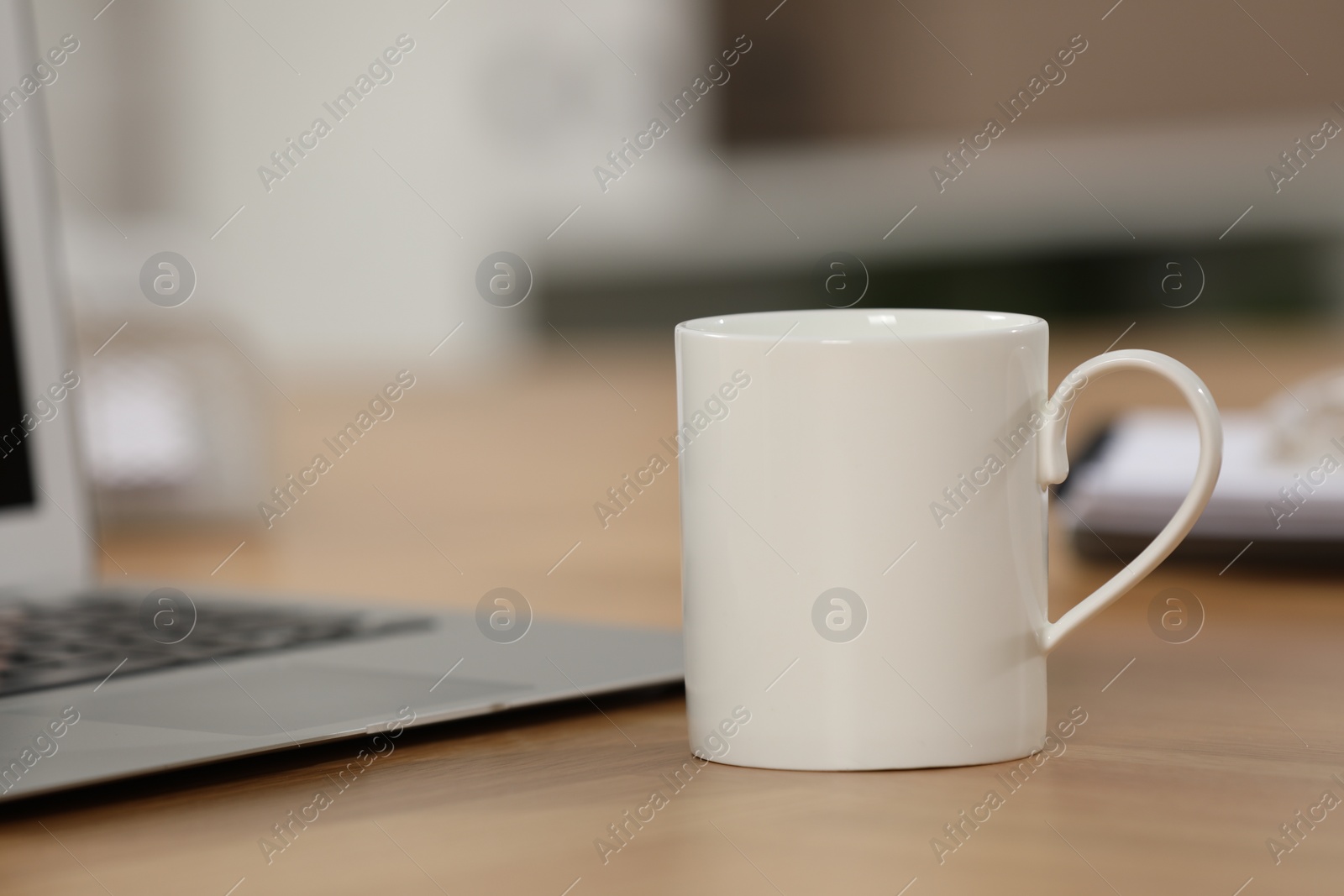 Photo of White ceramic mug and laptop on wooden table at workplace. Space for text