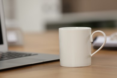 White ceramic mug and laptop on wooden table at workplace. Space for text