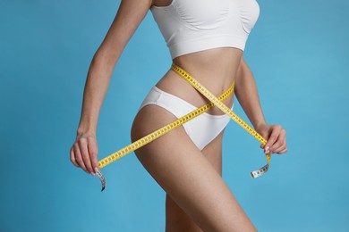 Young woman measuring waist with tape on light blue background, closeup