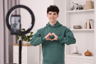 Photo of Smiling teenage blogger making heart gesture to his subscribers while streaming at home