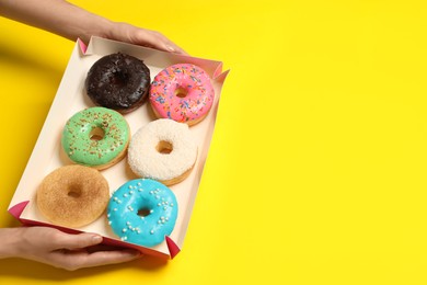 Woman with box of delicious donuts on yellow background, closeup. Space for text