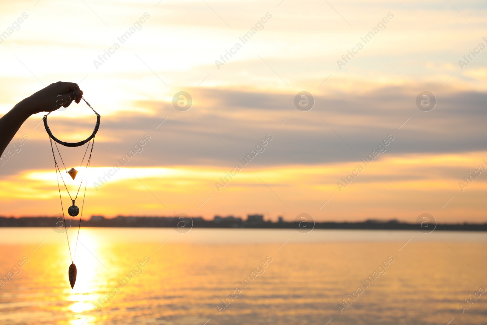 Photo of Young woman holding amulet near river at sunset, closeup view with space for text. Nature healing power