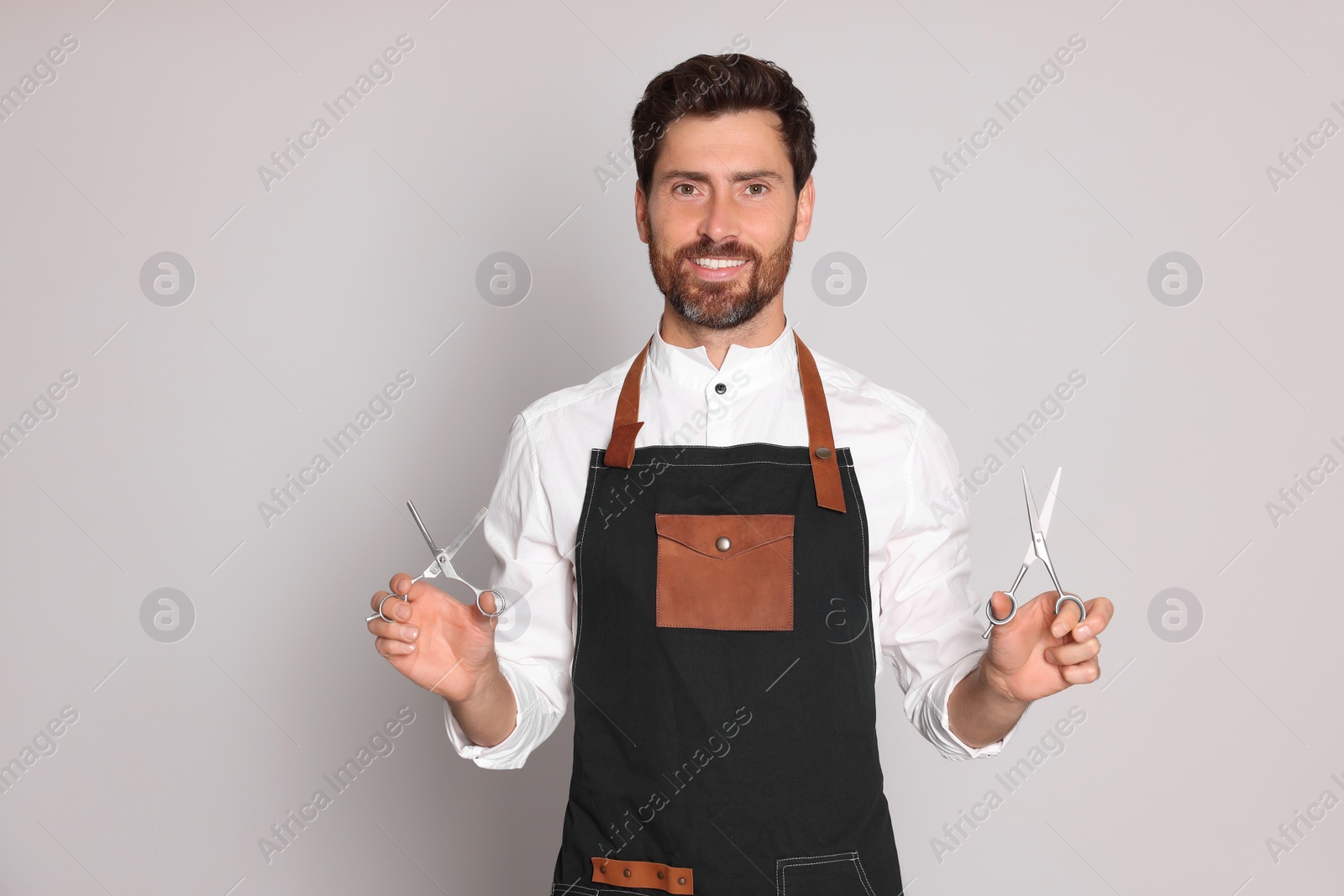 Photo of Smiling hairdresser wearing apron holding scissors on light grey background