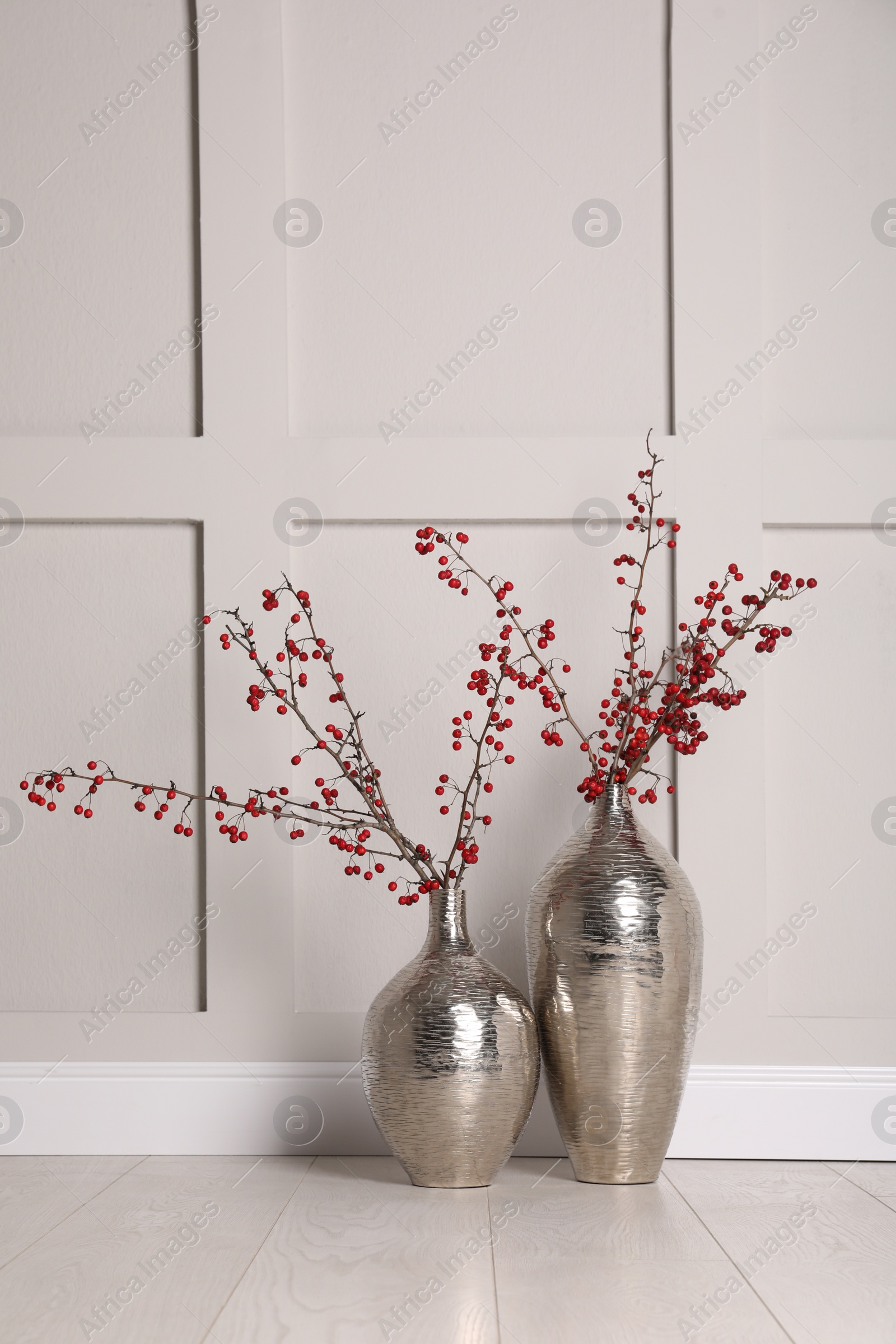 Photo of Hawthorn branches with red berries in vases near light  wall indoors