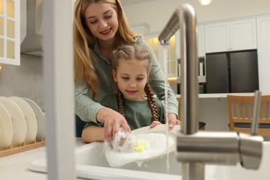 Photo of Mother and daughter washing plate above sink in kitchen, view from outside