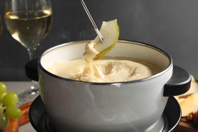 Photo of Dipping piece of pear into fondue pot with melted cheese at table on grey background, closeup