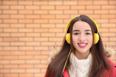 Photo of Beautiful young woman listening to music with headphones against brick wall. Space for text