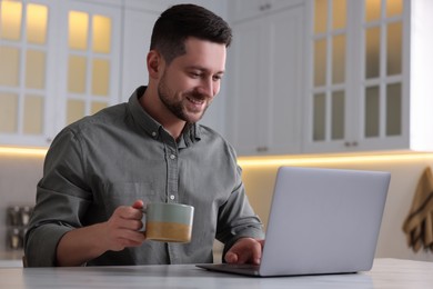 Photo of Happy man with cup of drink working on laptop at white marble table in kitchen