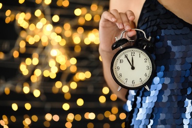 Photo of Woman holding alarm clock against blurred lights, closeup with space for text. New Year countdown