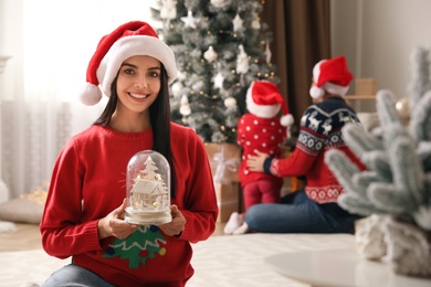 Photo of Woman in red Christmas sweater holding decorative snow globe at home