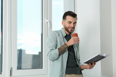 Photo of Handsome man with delicious smoothie reading book near window. Space for text