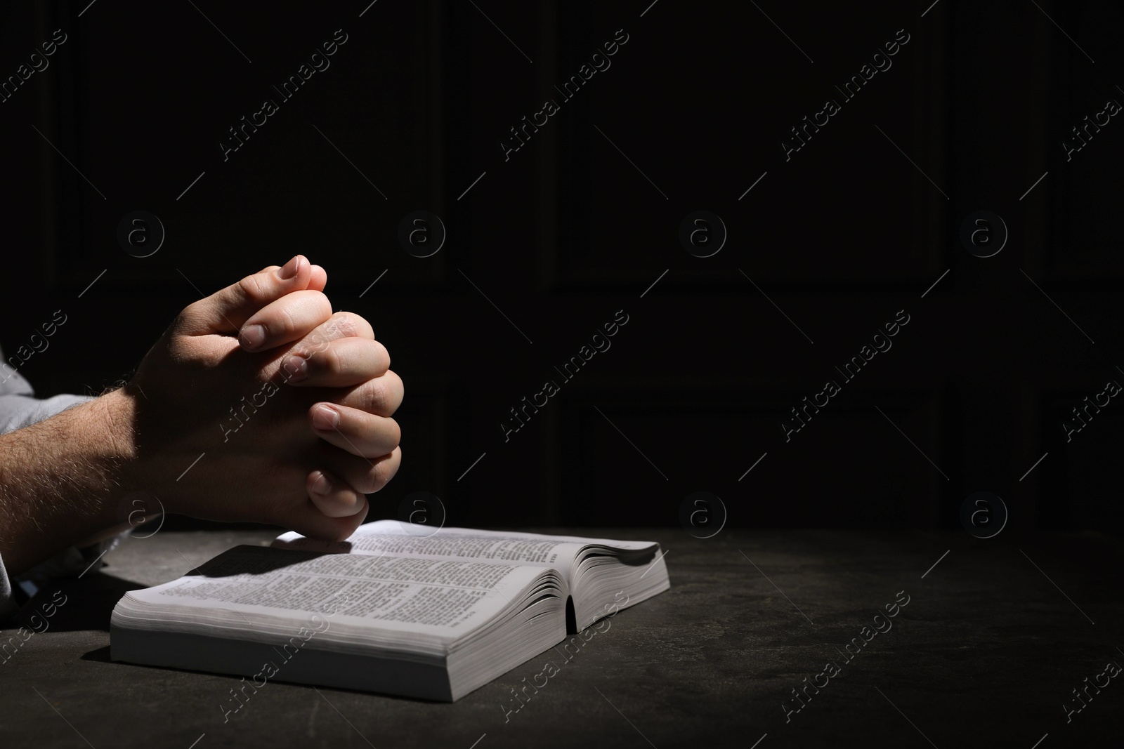 Photo of Religion. Christian man praying over Bible at table against black background, closeup. Space for text