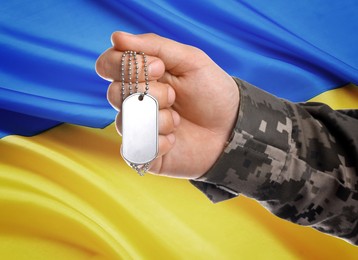 Closeup view of soldier in uniform holding military ID tag and Ukrainian flag on background