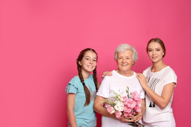 Photo of Happy sisters with their grandmother holding flowers on pink background