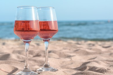 Photo of Glasses of tasty rose wine on sand near sea, space for text