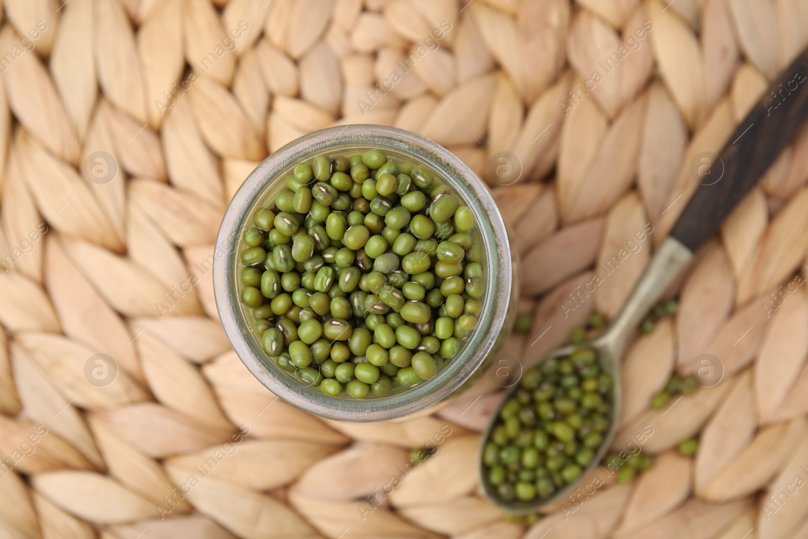 Photo of Glass jar of mung beans with spoon on wicker mat, flat lay