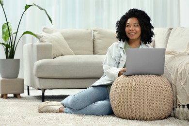 Photo of Happy young woman using laptop on pouf at home. Space for text