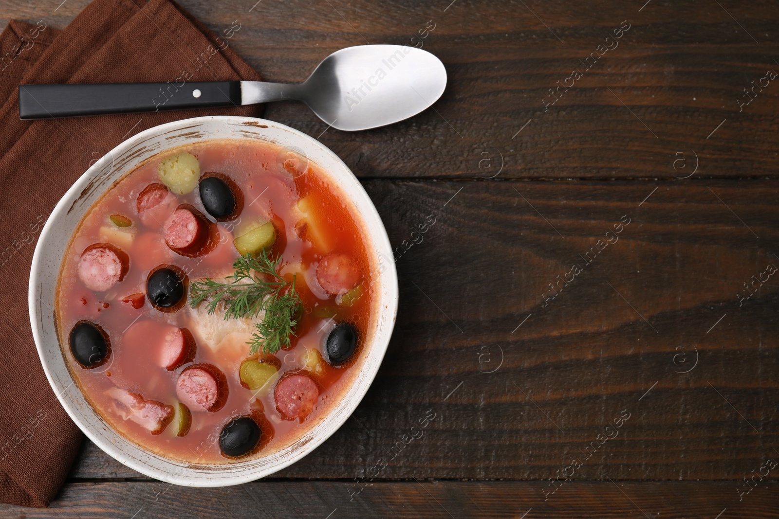 Photo of Meat solyanka soup with thin dry smoked sausages in bowl on wooden table, top view