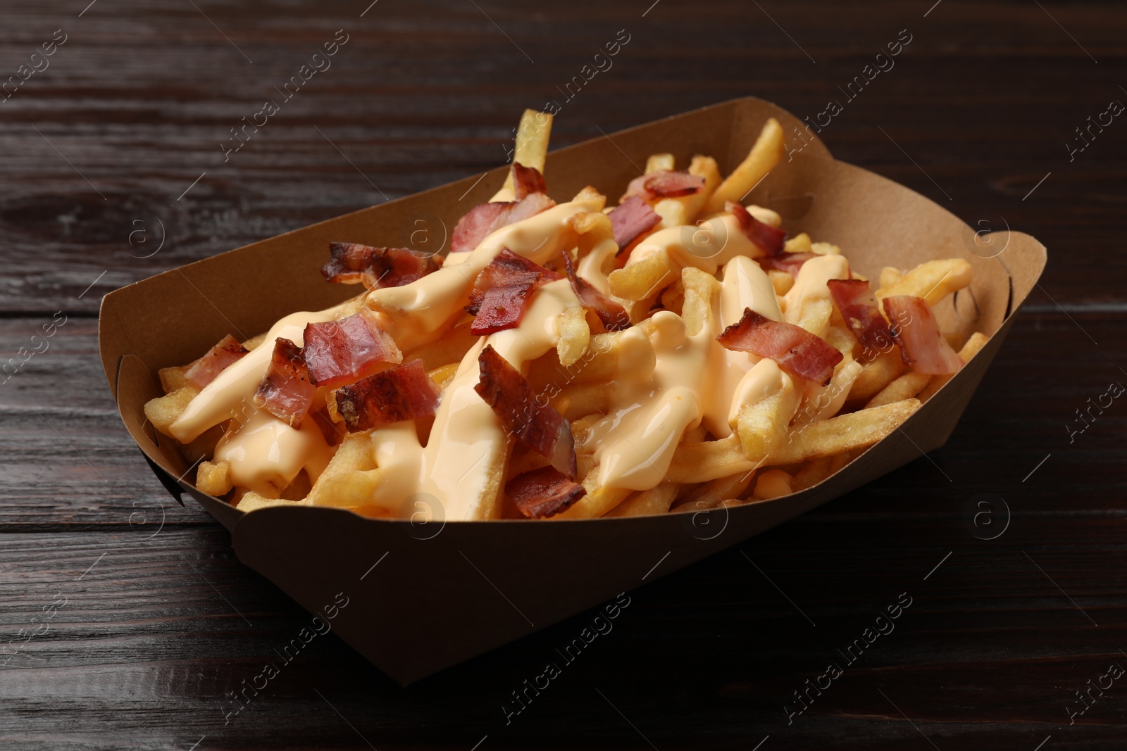 Photo of Tasty potato fries, cheese sauce and bacon in paper container on wooden table, closeup