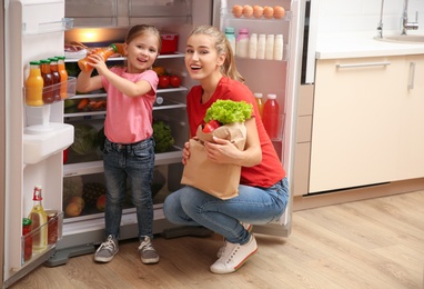 Photo of Young mother with daughter putting food into refrigerator at home after shopping