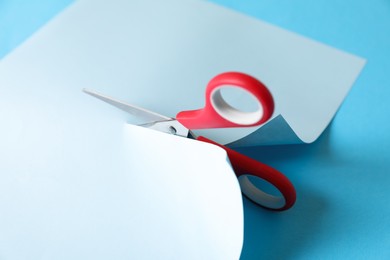 Red scissors cutting color paper on light blue background, closeup