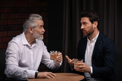 Photo of Men with glasses of whiskey talking at wooden table indoors