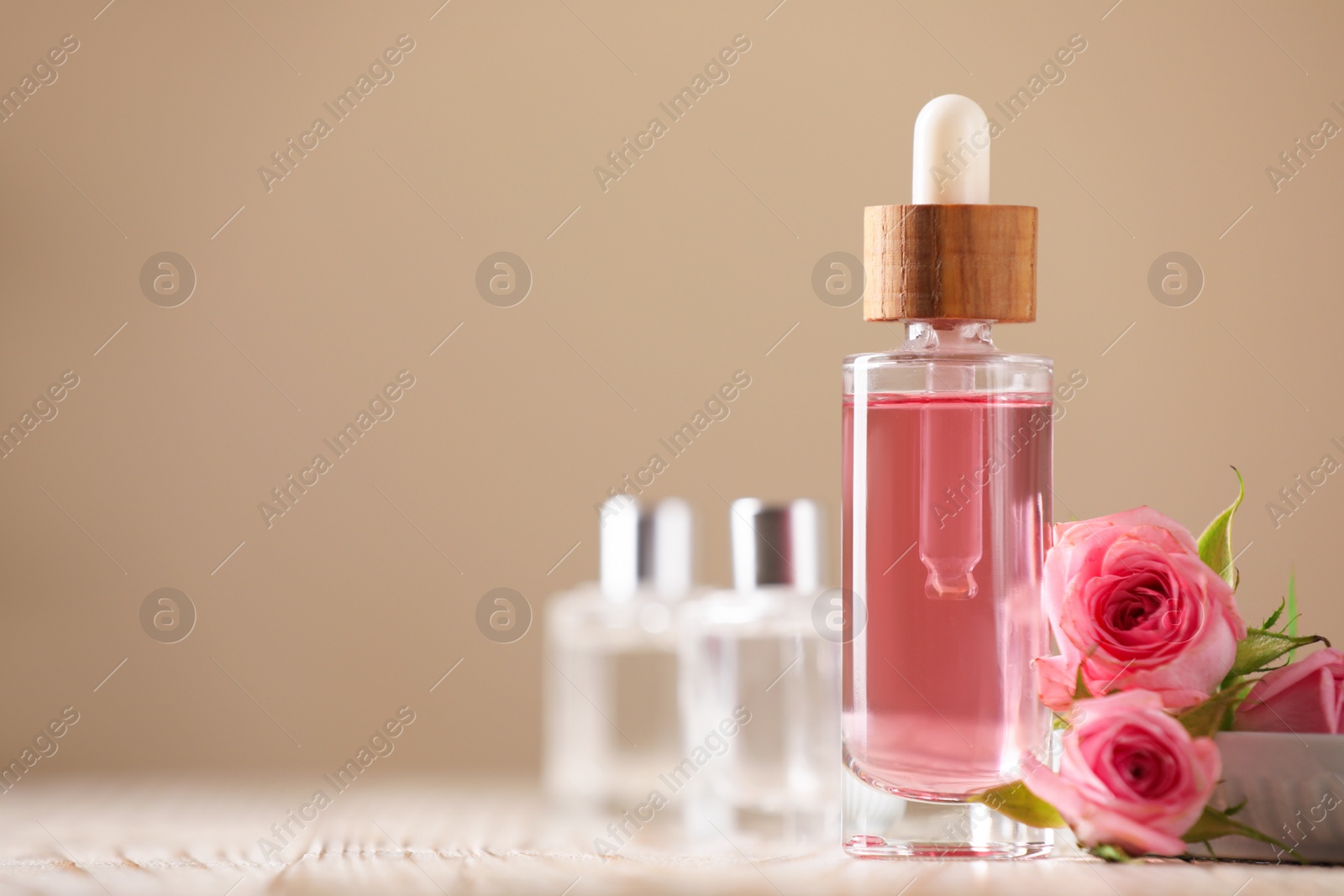 Photo of Bottle of essential rose oil and flowers on white wooden table against beige background, space for text