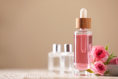 Photo of Bottle of essential rose oil and flowers on white wooden table against beige background, space for text
