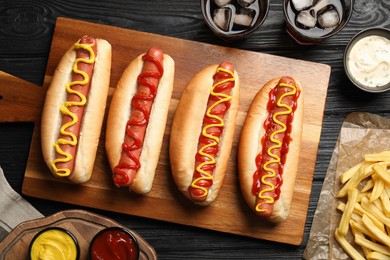 Photo of Delicious hot dogs with mustard, ketchup and potato fries on black wooden table, flat lay