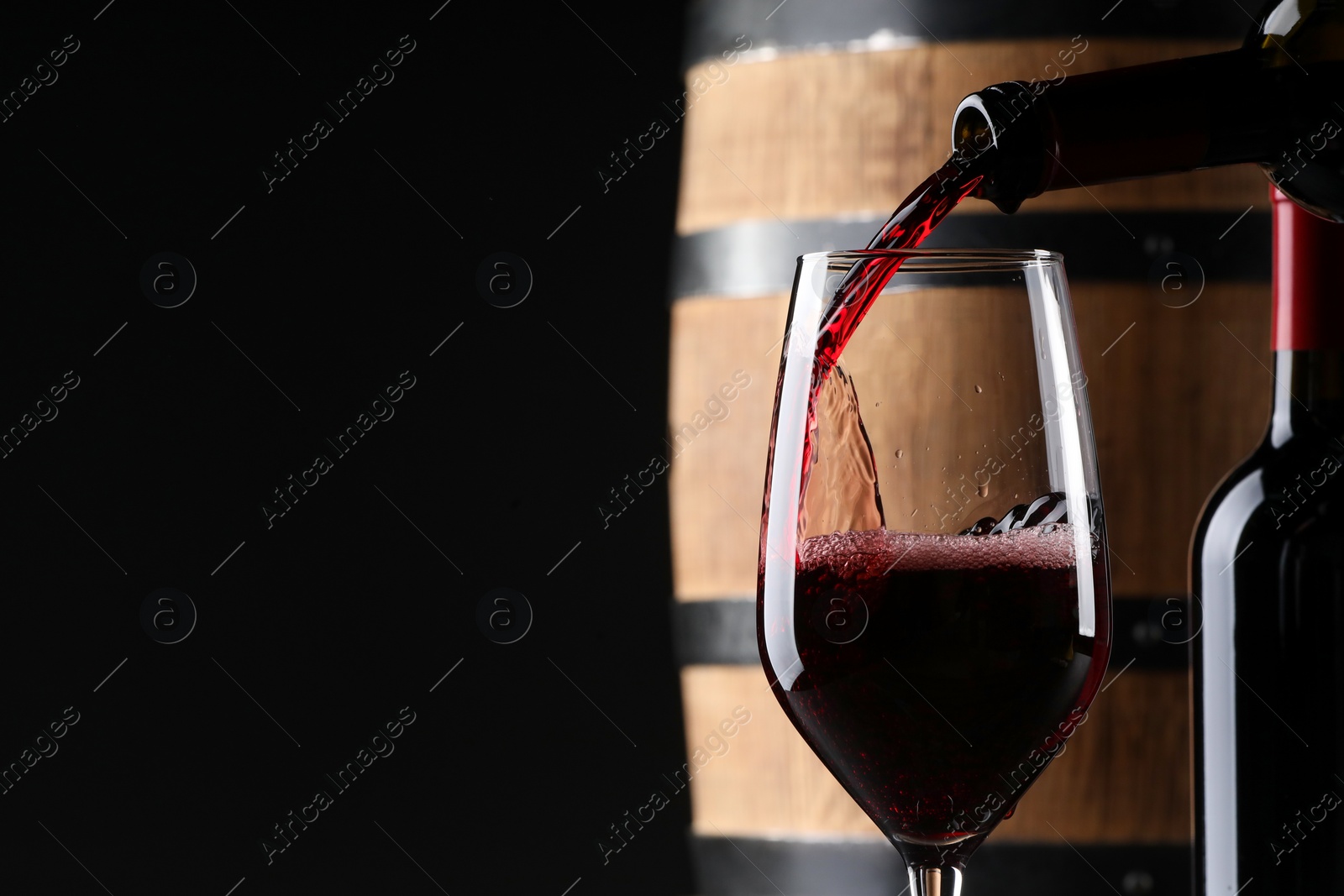 Photo of Pouring red wine from bottle into near wooden barrel against black background. Space for text