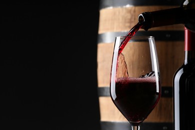 Pouring red wine from bottle into near wooden barrel against black background. Space for text