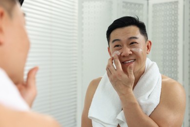 Handsome man applying cream onto his face near mirror indoors