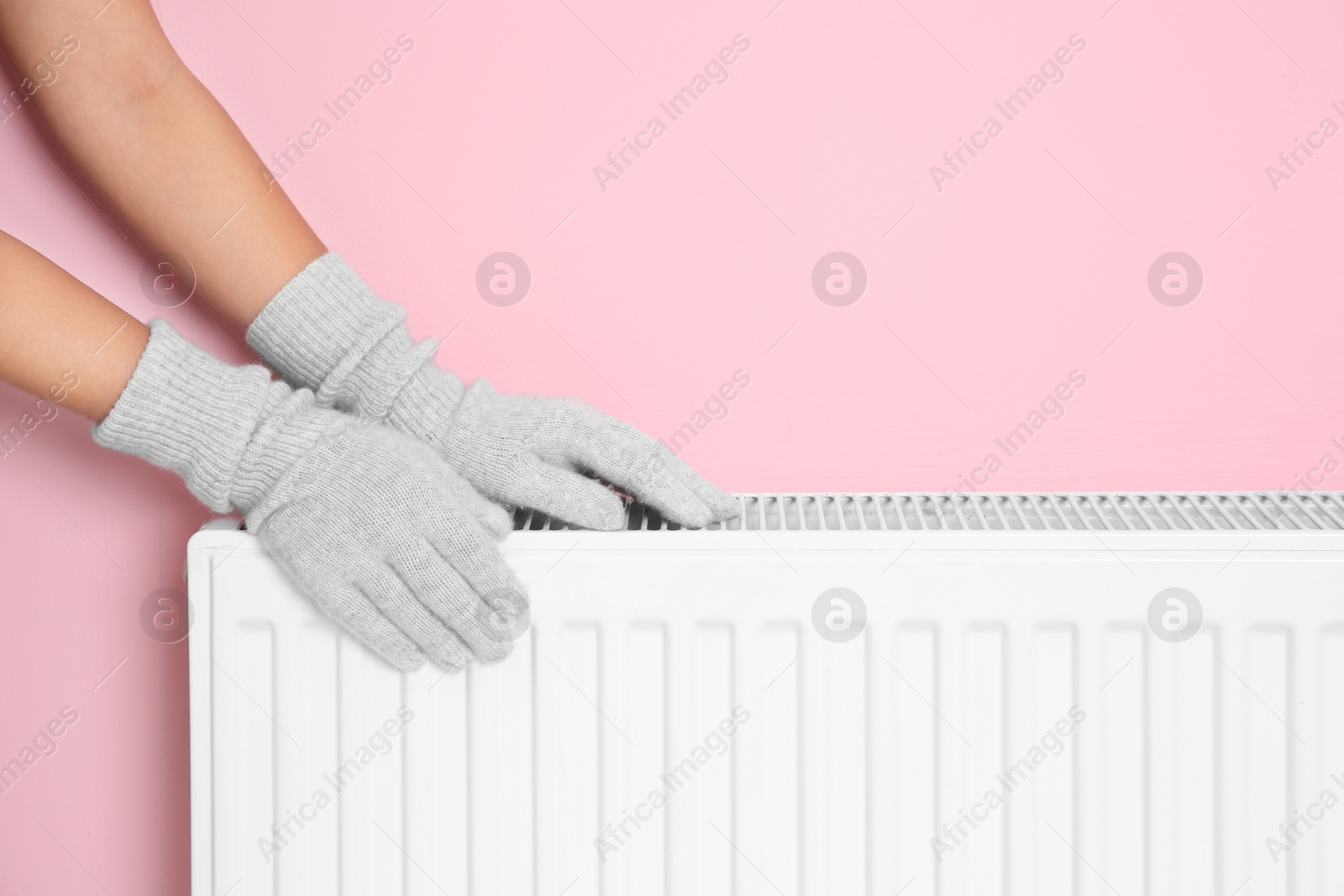 Photo of Woman in gloves warming hands on heating radiator near color wall