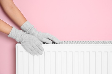 Woman in gloves warming hands on heating radiator near color wall