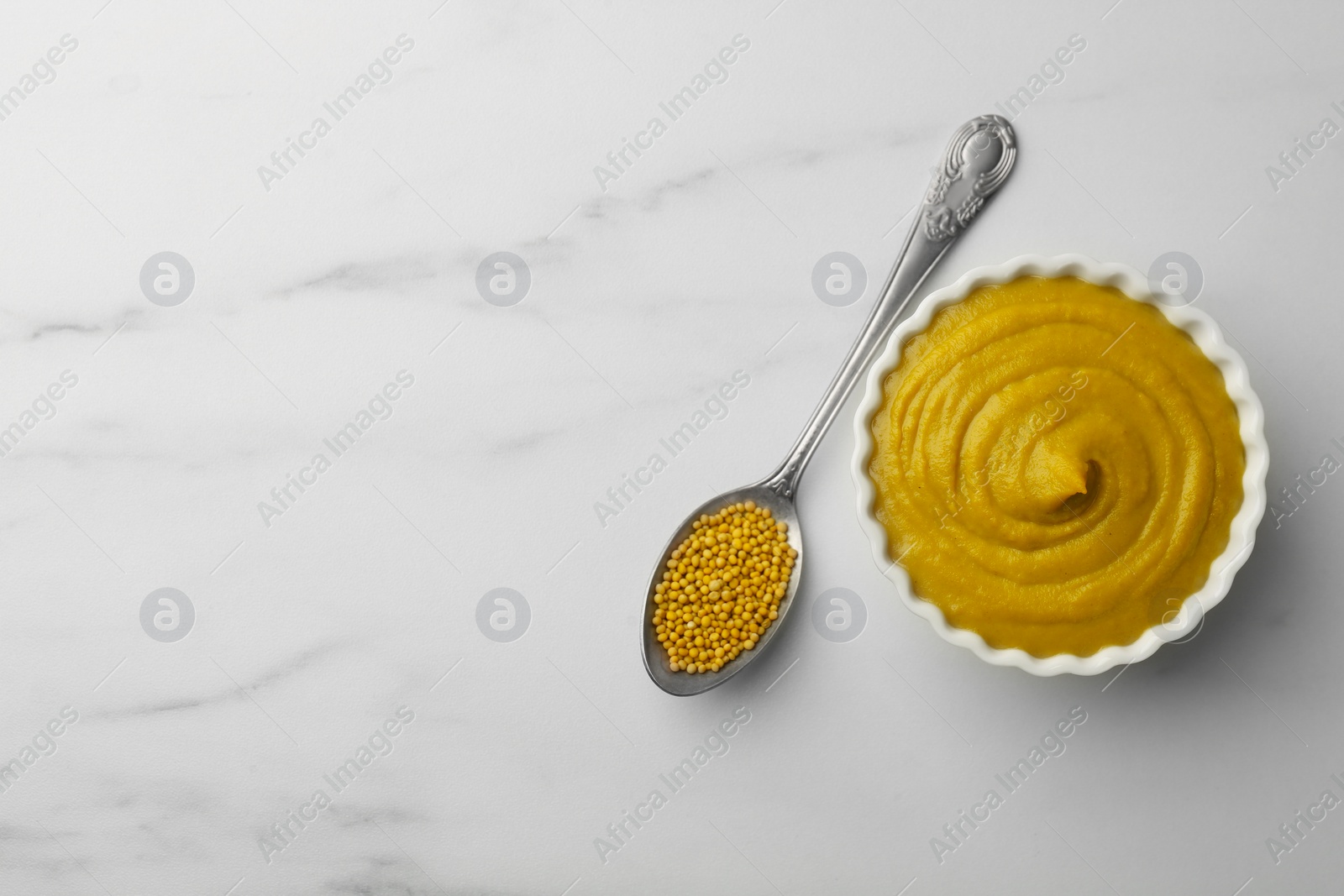 Photo of Tasty mustard sauce in bowl and spoon with dry seeds on white marble table, flat lay. Space for text